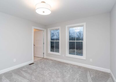 Empty gray bedroom with two large white-trimmed windows, an open door to a hallway, and a ceiling light fixture above.