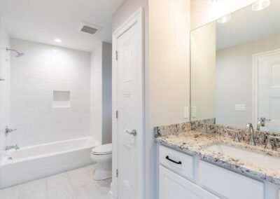 Bright, modern bathroom with a white-tiled shower and built-in niche, a toilet, and a white vanity with a granite countertop and chrome fixtures reflected in a large mirror.