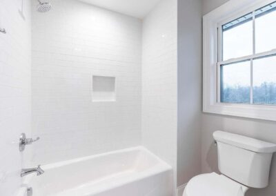 White tiled shower with built-in niche, chrome faucet, and a toilet beside a large window.