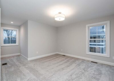 Empty room with light gray walls, white baseboards, and a cream carpet; two white-framed windows (left and right) and a central ceiling light fixture.