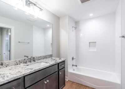 Dark wood vanity with speckled granite countertop, large mirror, and white-tiled bathtub/shower area.