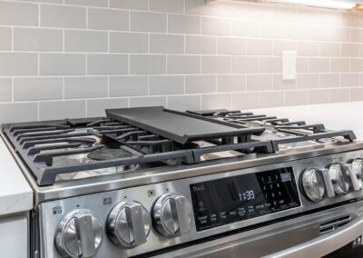 Stainless steel gas range with large grate over a flat grill insert, in a modern kitchen with gray subway tile backsplash.