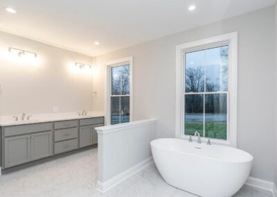 Modern bathroom with a double vanity, gray cabinets, and white countertops; sconces above mirror wall; freestanding oval tub by large window.