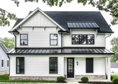 Front view of a two-story white house with a dark metal roof, black-framed windows, brick foundation, and a green lawn in the foreground.