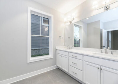 Bright modern bathroom with white double vanity, twin sinks, and large mirror above, flanked by wall-mounted lights; gray walls, white cabinetry, and a window overlooking the exterior.