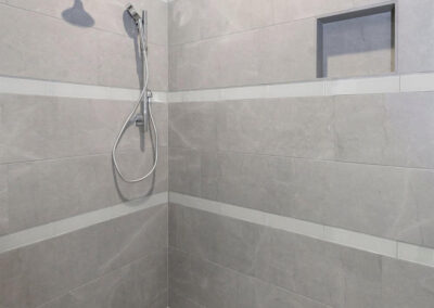 Modern gray tiled shower with fixed rain shower and handheld showerhead, built-in niche in wall, light grout lines and small square floor tiles.