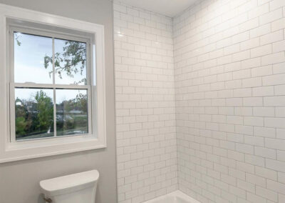 Small bathroom with white subway-tiled shower wall, white bathtub, and a white toilet beneath a window; gray walls and recessed ceiling lights visible.