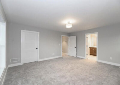 Empty gray living room with light carpet, large window on the left, two white doors, and an open doorway to a bathroom; ceiling light overhead.