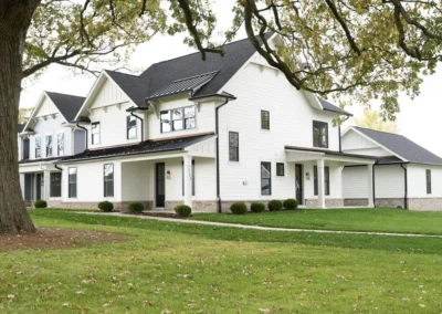 White multi‑story suburban house with a dark roof, covered porch, and manicured lawn framed by trees on a sunny day