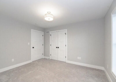 Bright empty bedroom with light gray walls, white trim, and beige carpet; features a ceiling light, a single door, a double closet, and a window along the right wall.