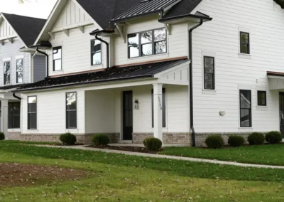 White two-story house with a black roof, front porch, and surrounding green lawn. Trees frame the scene above the home.