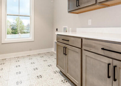 Modern kitchen corner: gray shaker cabinets, white countertop, and patterned tiled floor beneath a large window.